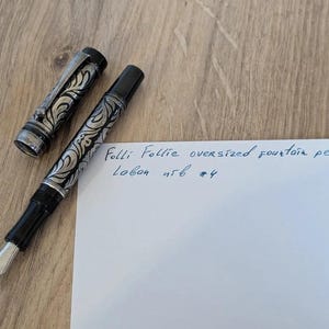以下が含まれることがあります： シルバーと黒のフローラルデザインの特大万年筆。キャップは横にあります。ペンは、青いインクで書かれた「Folli Follie oversized fountain pen Lobon 456 #4」というテキストが書かれた紙の隣にあります。