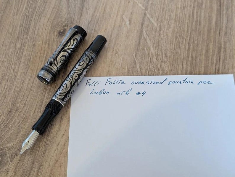 以下が含まれることがあります： シルバーと黒のフローラルデザインの特大万年筆。キャップは横にあります。ペンは、青いインクで書かれた「Folli Follie oversized fountain pen Lobon 456 #4」というテキストが書かれた紙の隣にあります。