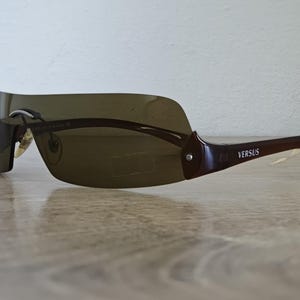 May include: Brown Versus sunglasses with a wraparound design. The sunglasses have a dark tinted lens and brown frames. The Versus logo is visible on the temple. The sunglasses are on a wooden surface.