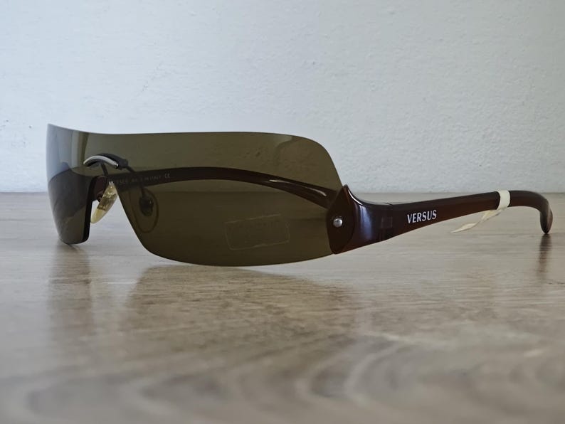 May include: Brown Versus sunglasses with a wraparound design. The sunglasses have a dark tinted lens and brown frames. The Versus logo is visible on the temple. The sunglasses are on a wooden surface.