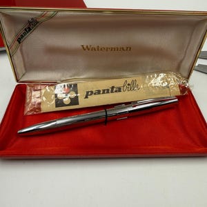 K&ouml;nnte beinhalten: Ein silberner Waterman-Stift in einem offenen roten Etui. Das Innere des Etuis ist beige mit dem Aufdruck "Waterman". Das Etui hat einen goldenen Rand und ein Band mit der Aufschrift "panta bille". Ein kleines Paket ist ebenfalls sichtbar.