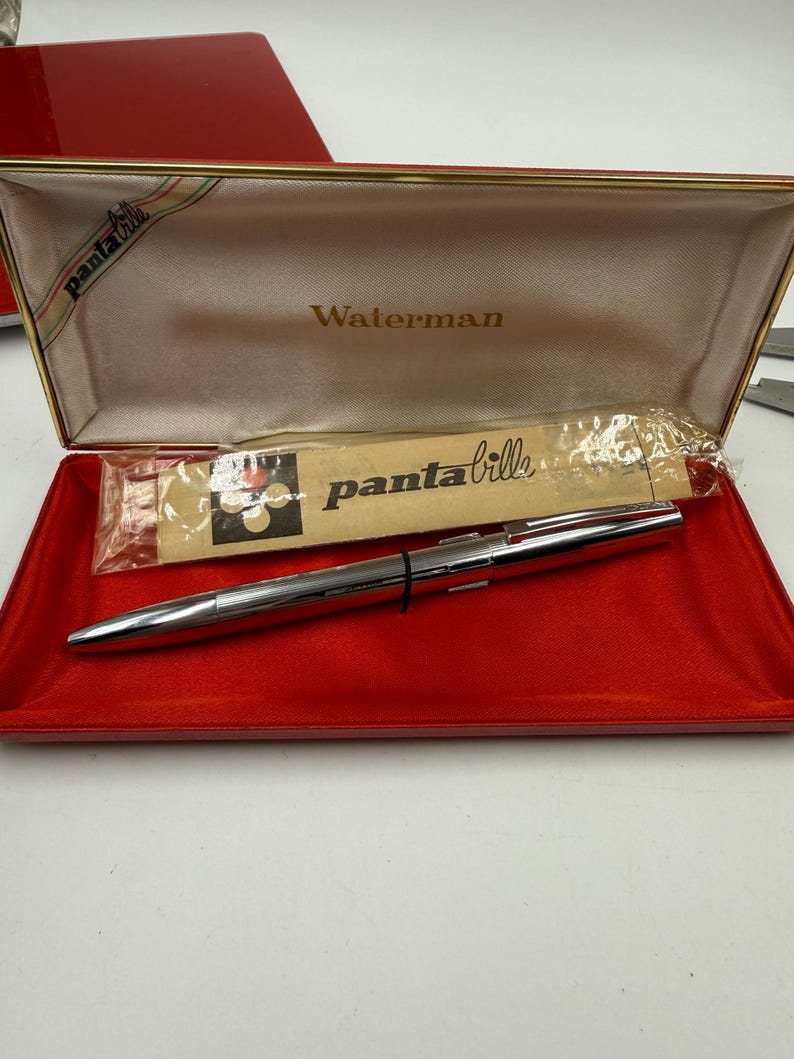 K&ouml;nnte beinhalten: Ein silberner Waterman-Stift in einem offenen roten Etui. Das Innere des Etuis ist beige mit dem Aufdruck "Waterman". Das Etui hat einen goldenen Rand und ein Band mit der Aufschrift "panta bille". Ein kleines Paket ist ebenfalls sichtbar.