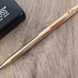 Könnte beinhalten: Ein goldfarbener Cross-Stift mit schwarzer Spitze und schwarzem Etui. Der Stift liegt auf einer Holzoberfläche. Auf dem schwarzen Etui steht "CROSS" und "SINCE 1846".