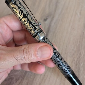 以下が含まれることがあります： 銀と黒の万年筆。装飾的な花柄のデザイン。ペンはシルバーのクリップと先端と上部に黒のアクセントが付いています。手で持っています。