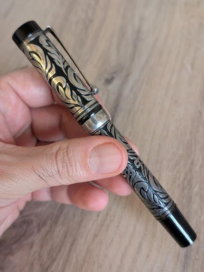以下が含まれることがあります： 銀と黒の万年筆。装飾的な花柄のデザイン。ペンはシルバーのクリップと先端と上部に黒のアクセントが付いています。手で持っています。