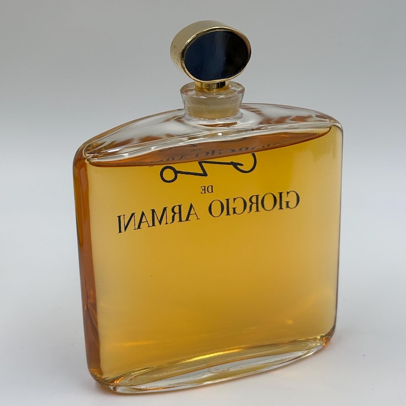 May include: A clear glass perfume bottle with a gold cap and dark blue center. The bottle contains a golden-colored liquid and features the text "GIORGIO ARMANI DE" in black lettering. The bottle has a unique, angular shape.