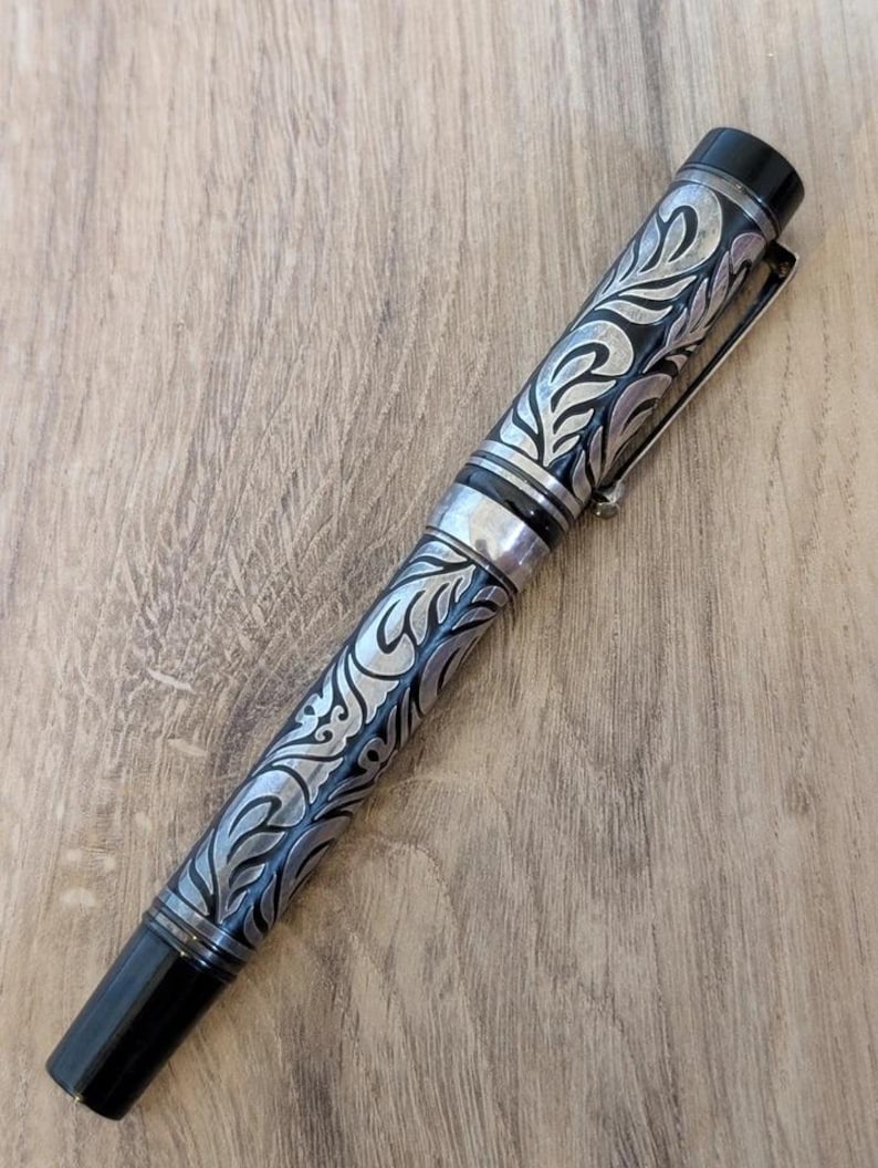 以下が含まれることがあります： 銀と黒の万年筆。装飾的なフローラルデザイン。ペンは黒いキャップとペン先、シルバーのクリップ付き。ペンの本体には、詳細な渦巻き模様が刻まれています。