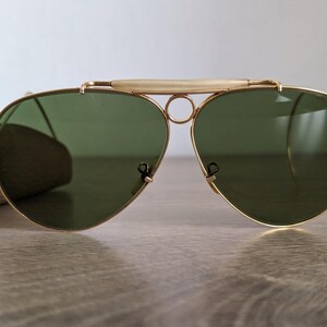 K&ouml;nnte beinhalten: Goldgerahmte Aviator-Sonnenbrille mit gr&uuml;nen Gl&auml;sern. Die Sonnenbrille hat einen Doppelsteg und einen Stirnb&uuml;gel. Die Gl&auml;ser sind tropfenf&ouml;rmig. Die Sonnenbrille liegt auf einer Holzoberfl&auml;che.