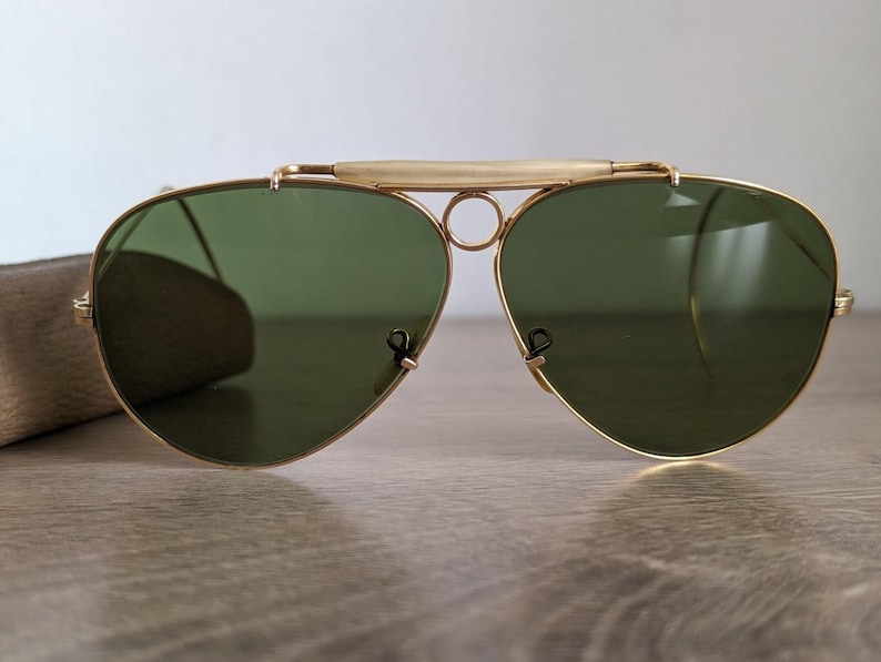 K&ouml;nnte beinhalten: Goldgerahmte Aviator-Sonnenbrille mit gr&uuml;nen Gl&auml;sern. Die Sonnenbrille hat einen Doppelsteg und einen Stirnb&uuml;gel. Die Gl&auml;ser sind tropfenf&ouml;rmig. Die Sonnenbrille liegt auf einer Holzoberfl&auml;che.