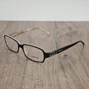May include: Rectangular spectacles with a black front frame and light beige inner rim. The temples are black with a silver-toned metallic accent featuring a Greek key pattern and the word VERSACE. The word VERSACE is also printed on the lens.