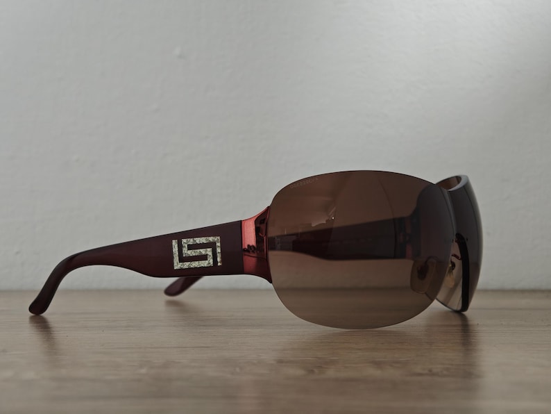 May include: Brown-tinted sunglasses with a dark burgundy frame. The sunglasses feature a large, rounded lens design and a decorative emblem with a square shape and embedded crystals on the temple.