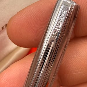 K&ouml;nnte beinhalten: Nahaufnahme eines silbernen Stifts mit dem Wort "WATERMAN" auf der Kappe eingraviert. Der Stift hat vertikale Linien entlang seines K&ouml;rpers. Der Stift wird in der Hand einer Person gehalten.