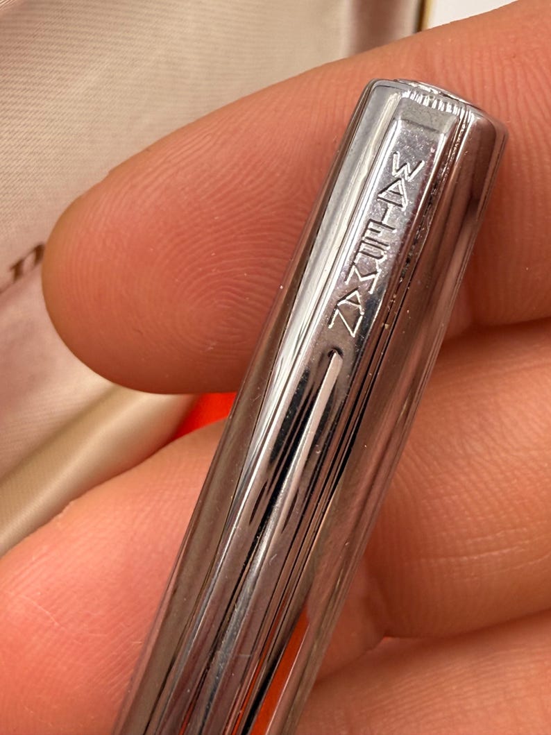 K&ouml;nnte beinhalten: Nahaufnahme eines silbernen Stifts mit dem Wort "WATERMAN" auf der Kappe eingraviert. Der Stift hat vertikale Linien entlang seines K&ouml;rpers. Der Stift wird in der Hand einer Person gehalten.