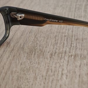 Op de afbeelding: Close-up van een bril met een donkergroen montuur en bruine pootjes. De tekst "Candidate 4.0" staat op de poot. De bril ligt op een lichtbruin houten oppervlak.