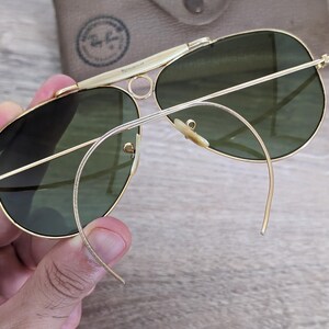 K&ouml;nnte beinhalten: Goldfarbene Aviator-Sonnenbrille mit gr&uuml;nen Gl&auml;sern. Die Sonnenbrille hat einen Doppelsteg und gebogene B&uuml;gel. Im Hintergrund ist ein beiges Etui mit Logo zu sehen. Die Sonnenbrille wird von einer Hand gehalten.
