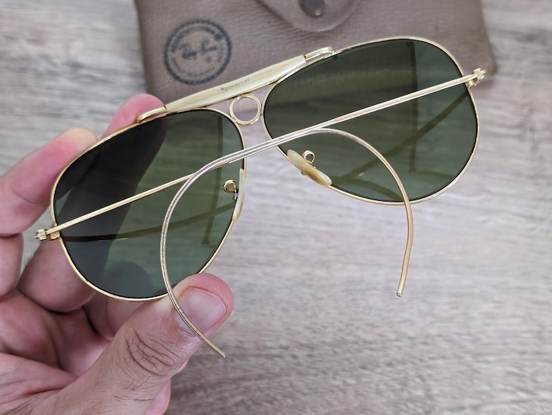K&ouml;nnte beinhalten: Goldfarbene Aviator-Sonnenbrille mit gr&uuml;nen Gl&auml;sern. Die Sonnenbrille hat einen Doppelsteg und gebogene B&uuml;gel. Im Hintergrund ist ein beiges Etui mit Logo zu sehen. Die Sonnenbrille wird von einer Hand gehalten.