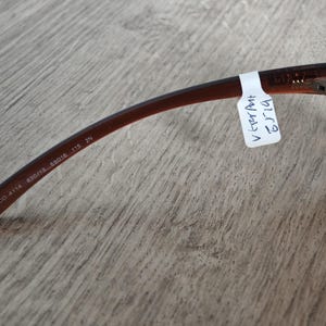 Puede incluir: Gafas de color marr&oacute;n con una patilla curvada. La patilla tiene el texto "MCO 4114 830/13 59016 115 2N". Una etiqueta blanca con texto manuscrito est&aacute; adherida a la patilla. Las gafas est&aacute;n sobre una superficie de madera.