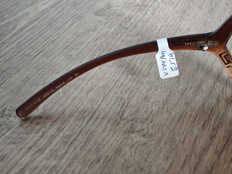 Puede incluir: Gafas de color marr&oacute;n con una patilla curvada. La patilla tiene el texto "MCO 4114 830/13 59016 115 2N". Una etiqueta blanca con texto manuscrito est&aacute; adherida a la patilla. Las gafas est&aacute;n sobre una superficie de madera.