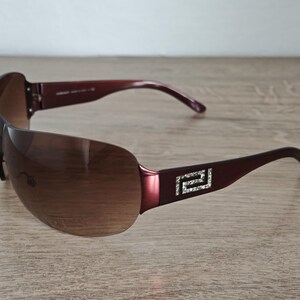 May include: Brown rimless sunglasses with a dark burgundy frame and arms. The arms feature a silver and crystal design. The lenses are a gradient brown color. The sunglasses are resting on a wooden surface.