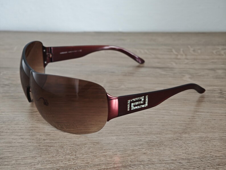 May include: Brown rimless sunglasses with a dark burgundy frame and arms. The arms feature a silver and crystal design. The lenses are a gradient brown color. The sunglasses are resting on a wooden surface.