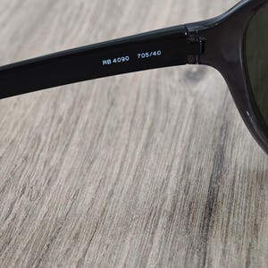 Puede incluir: Primer plano de gafas de sol negras con el texto "RB 4090 705/40" impreso en blanco en la patilla. Las gafas de sol est&aacute;n sobre una superficie de madera marr&oacute;n claro.