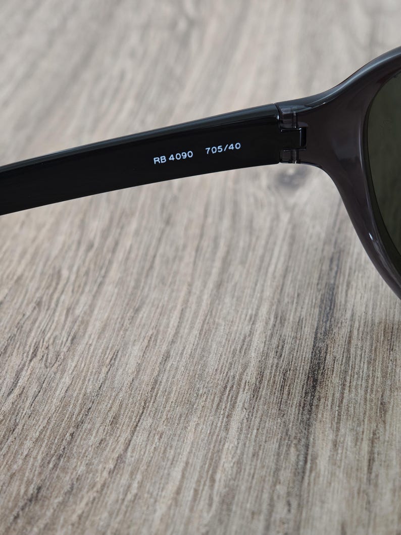 Puede incluir: Primer plano de gafas de sol negras con el texto "RB 4090 705/40" impreso en blanco en la patilla. Las gafas de sol est&aacute;n sobre una superficie de madera marr&oacute;n claro.