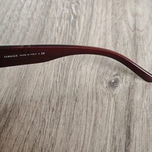 May include: Close-up of a burgundy-colored eyeglass arm. The text "VERSACE MADE IN ITALY A CE" is printed in white on the arm. The arm curves gently, with a smooth, glossy finish.
