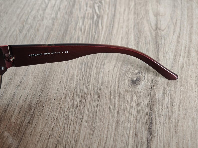 May include: Close-up of a burgundy-colored eyeglass arm. The text "VERSACE MADE IN ITALY A CE" is printed in white on the arm. The arm curves gently, with a smooth, glossy finish.