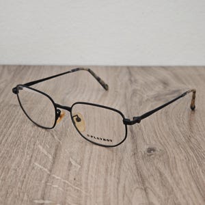 May include: Black metal eyeglasses with a thin frame and a hexagonal shape. The temples feature a brown and black tortoiseshell pattern at the tips. The right lens has the word PLAYBOY printed in black text. The nose pads are a light tan color.