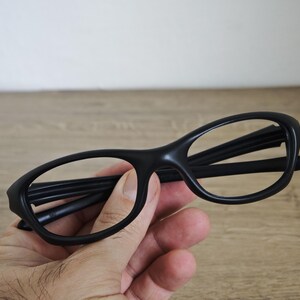 May include: Black eyeglasses with a rounded frame. The glasses are held in a hand against a wooden background. The frames are thick and the lenses are clear.