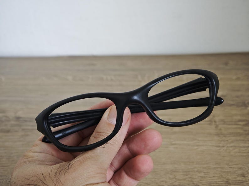 May include: Black eyeglasses with a rounded frame. The glasses are held in a hand against a wooden background. The frames are thick and the lenses are clear.