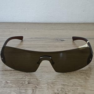 May include: Brown-tinted sunglasses with a curved, rimless design. The frames are dark brown with a bridge and temples. The lenses are a dark brown color. The sunglasses are resting on a wooden surface.
