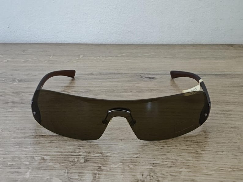 May include: Brown-tinted sunglasses with a curved, rimless design. The frames are dark brown with a bridge and temples. The lenses are a dark brown color. The sunglasses are resting on a wooden surface.