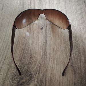 May include: Brown sunglasses with a curved, rimless design. The lenses have a gradient tint, transitioning from a darker shade at the top to a lighter shade at the bottom. The temples are a dark brown color.