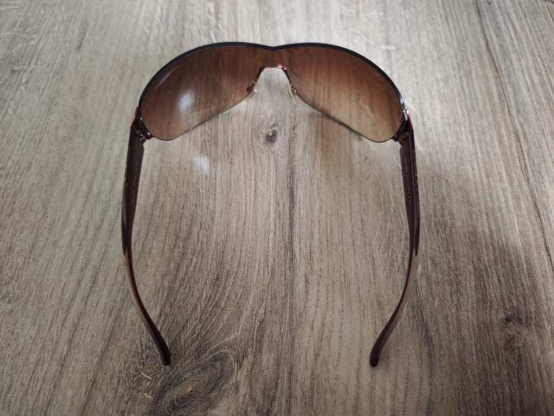 May include: Brown sunglasses with a curved, rimless design. The lenses have a gradient tint, transitioning from a darker shade at the top to a lighter shade at the bottom. The temples are a dark brown color.
