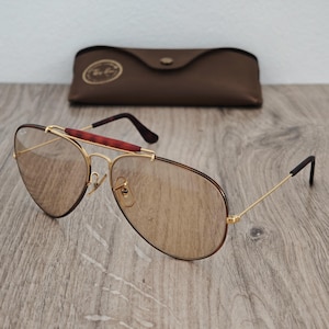 K&ouml;nnte beinhalten: Goldfarbene Pilotenbrille mit braunen Gl&auml;sern und einem braunen Lederetui. Die Sonnenbrille hat einen rotbraunen Steg und dunkelbraune B&uuml;gel. Das Etui hat ein goldenes Emblem.
