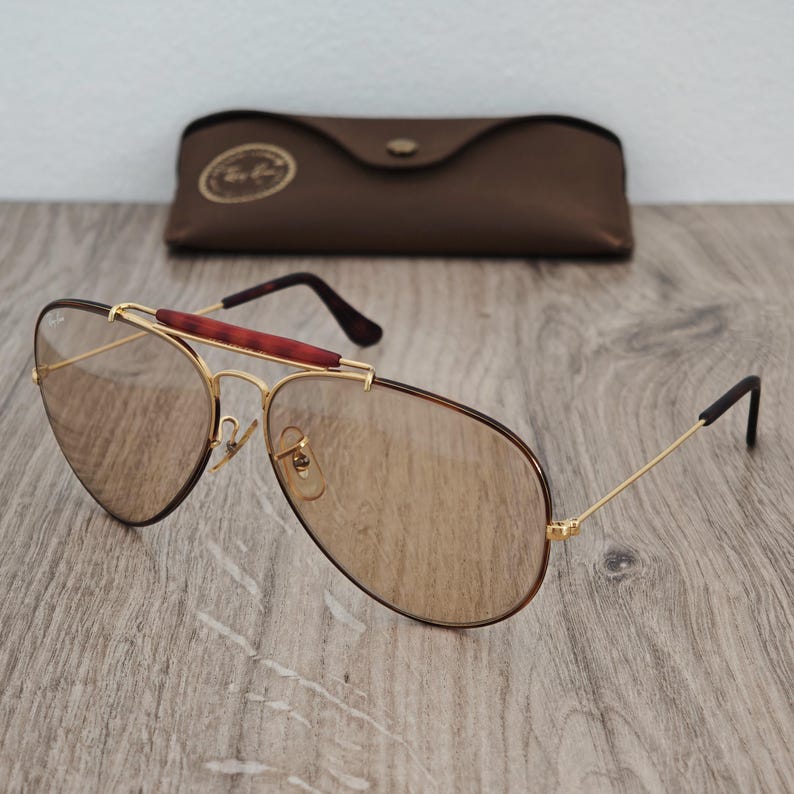 K&ouml;nnte beinhalten: Goldfarbene Pilotenbrille mit braunen Gl&auml;sern und einem braunen Lederetui. Die Sonnenbrille hat einen rotbraunen Steg und dunkelbraune B&uuml;gel. Das Etui hat ein goldenes Emblem.