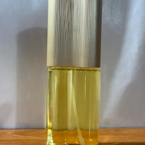 Puede incluir: Un frasco de perfume vintage con una tapa dorada y acanalada y un cuerpo de vidrio transparente. El frasco contiene un líquido amarillo. El frasco es rectangular con una base redondeada y un pulverizador.