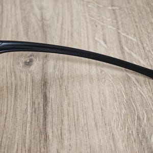 May include: Close-up of a black eyeglass arm. The arm is curved and tapers to a rounded tip. The frame is made of a smooth, matte material. The background is a light brown wooden surface.