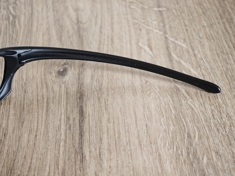 May include: Close-up of a black eyeglass arm. The arm is curved and tapers to a rounded tip. The frame is made of a smooth, matte material. The background is a light brown wooden surface.