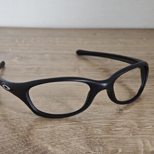 May include: Black eyeglasses with a curved frame and oval lenses. The glasses have a sleek, modern design and a small silver logo on the temple. The frames are made of a matte material.
