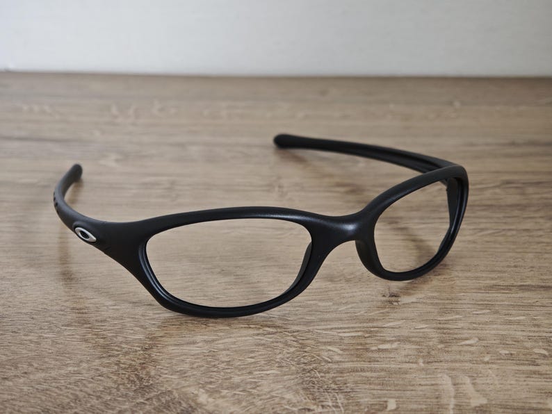 May include: Black eyeglasses with a curved frame and oval lenses. The glasses have a sleek, modern design and a small silver logo on the temple. The frames are made of a matte material.