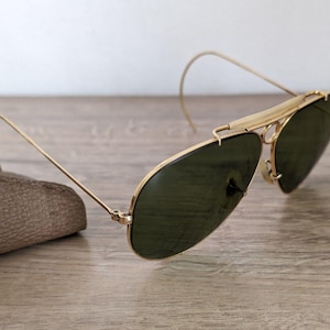 K&ouml;nnte beinhalten: Goldfarbene Aviator-Sonnenbrille mit dunkelgr&uuml;nen Gl&auml;sern. Die Sonnenbrille hat ein klassisches Design mit Doppelsteg und gebogenen B&uuml;geln. Die Gl&auml;ser sind oval und der Rahmen ist goldfarben gl&auml;nzend. Die Sonnenbrille liegt auf einer Holzoberfl&auml;che.