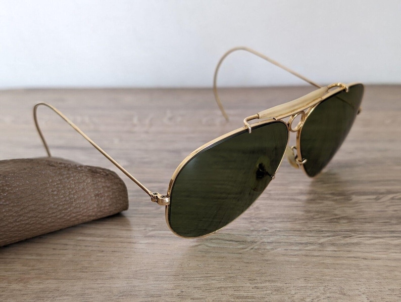 K&ouml;nnte beinhalten: Goldfarbene Aviator-Sonnenbrille mit dunkelgr&uuml;nen Gl&auml;sern. Die Sonnenbrille hat ein klassisches Design mit Doppelsteg und gebogenen B&uuml;geln. Die Gl&auml;ser sind oval und der Rahmen ist goldfarben gl&auml;nzend. Die Sonnenbrille liegt auf einer Holzoberfl&auml;che.