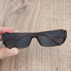 Puede incluir: Gafas de sol rectangulares con montura de pl&aacute;stico marr&oacute;n oscuro transl&uacute;cido y lentes oscuras. Las patillas est&aacute;n plegadas hacia adentro, mostrando el dise&ntilde;o estilizado de la montura. El marco presenta un acabado liso con un tono de color uniforme.