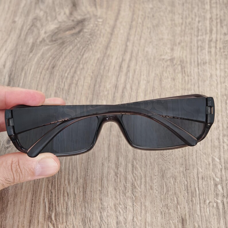 Puede incluir: Gafas de sol rectangulares con montura de pl&aacute;stico marr&oacute;n oscuro transl&uacute;cido y lentes oscuras. Las patillas est&aacute;n plegadas hacia adentro, mostrando el dise&ntilde;o estilizado de la montura. El marco presenta un acabado liso con un tono de color uniforme.
