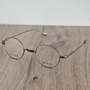 May include: Bronze-coloured, round eyeglasses with slender frames. The glasses feature a classic design, with the text "100% TITANIUM" on one lens and the signature "John Lennon" on the other. They are displayed on a wooden surface.