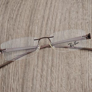 Puede incluir: Gafas sin montura con montura de metal marrón y lentes transparentes. El logotipo de Ray-Ban es visible en la patilla. Las gafas son de titanio.