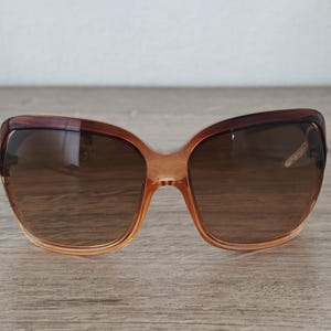 Puede incluir: Gafas de sol degradadas marrones con montura en forma de mariposa. Las lentes son de color marr&oacute;n oscuro y la montura pasa de un naranja claro a un marr&oacute;n m&aacute;s oscuro en la parte superior. Las gafas de sol est&aacute;n sobre una superficie de madera.