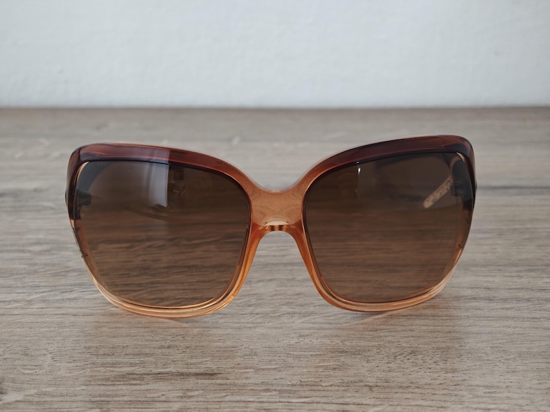 Puede incluir: Gafas de sol degradadas marrones con montura en forma de mariposa. Las lentes son de color marr&oacute;n oscuro y la montura pasa de un naranja claro a un marr&oacute;n m&aacute;s oscuro en la parte superior. Las gafas de sol est&aacute;n sobre una superficie de madera.